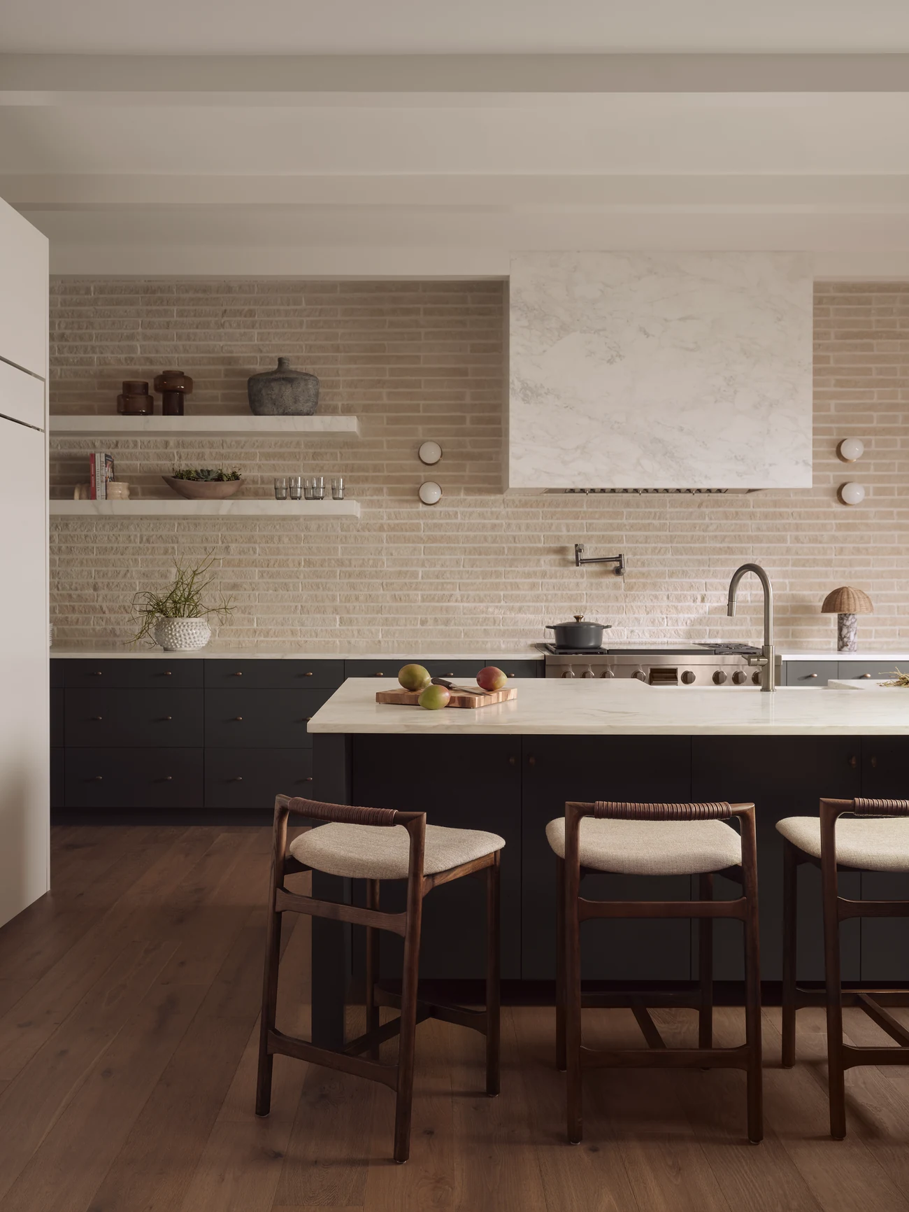 A kitchen with a stone backsplash, marble range hood and walnut counter stools
